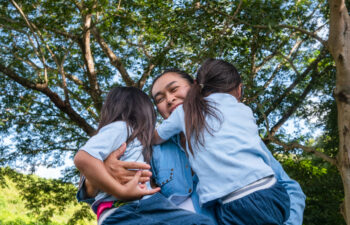 Two sibling little girls with mom laughing and hugging each other on warm and sunny summer day in the garden. Young girls with her mother spending day in park.