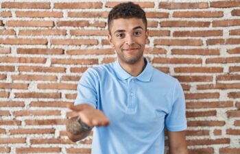 Brazilian young man standing over brick wall smiling cheerful offering palm hand giving assistance and acceptance.
